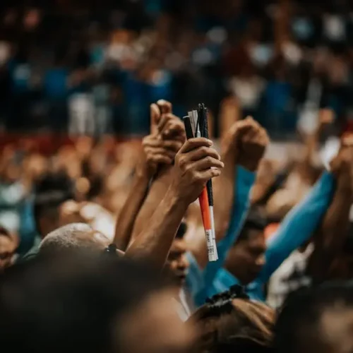 A crowd of people raising their hands with pens and fists in a large indoor venue.